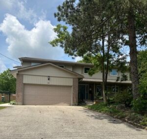 A beige house with a garage and multiple units on the second floor with green trees on the lawn out front and a stone driveway leading to the garage and front.