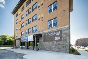 An orange brick 5 floor apartment building with a brown sign that reads 10 Shelldale in white letters. A blue and white unreadable signs hangs on the front entrance. 