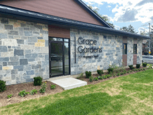 A grey stone and red sided single story building with the words Grace Gardens in black lettering next to a glass window entrance.