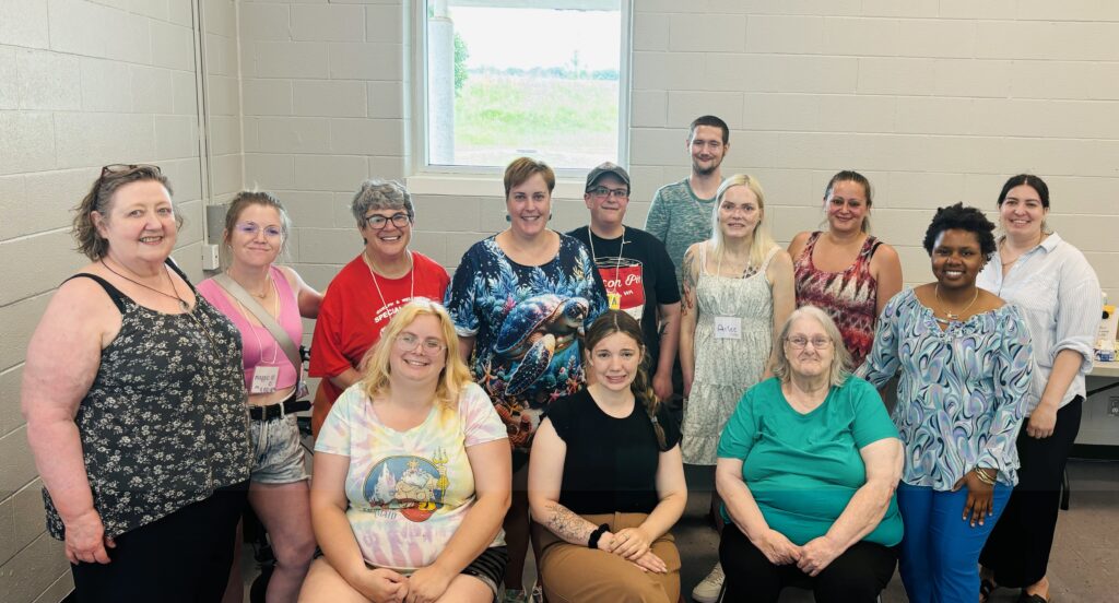 A group photo in front of a white wall with a window with 13 people facing forward smiling. 