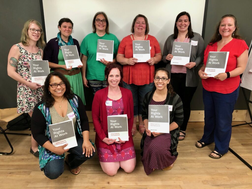 A group photo in front of 9 people facing forward smiling. Each person is holding a gray and white document that says 'your rights at work'. 